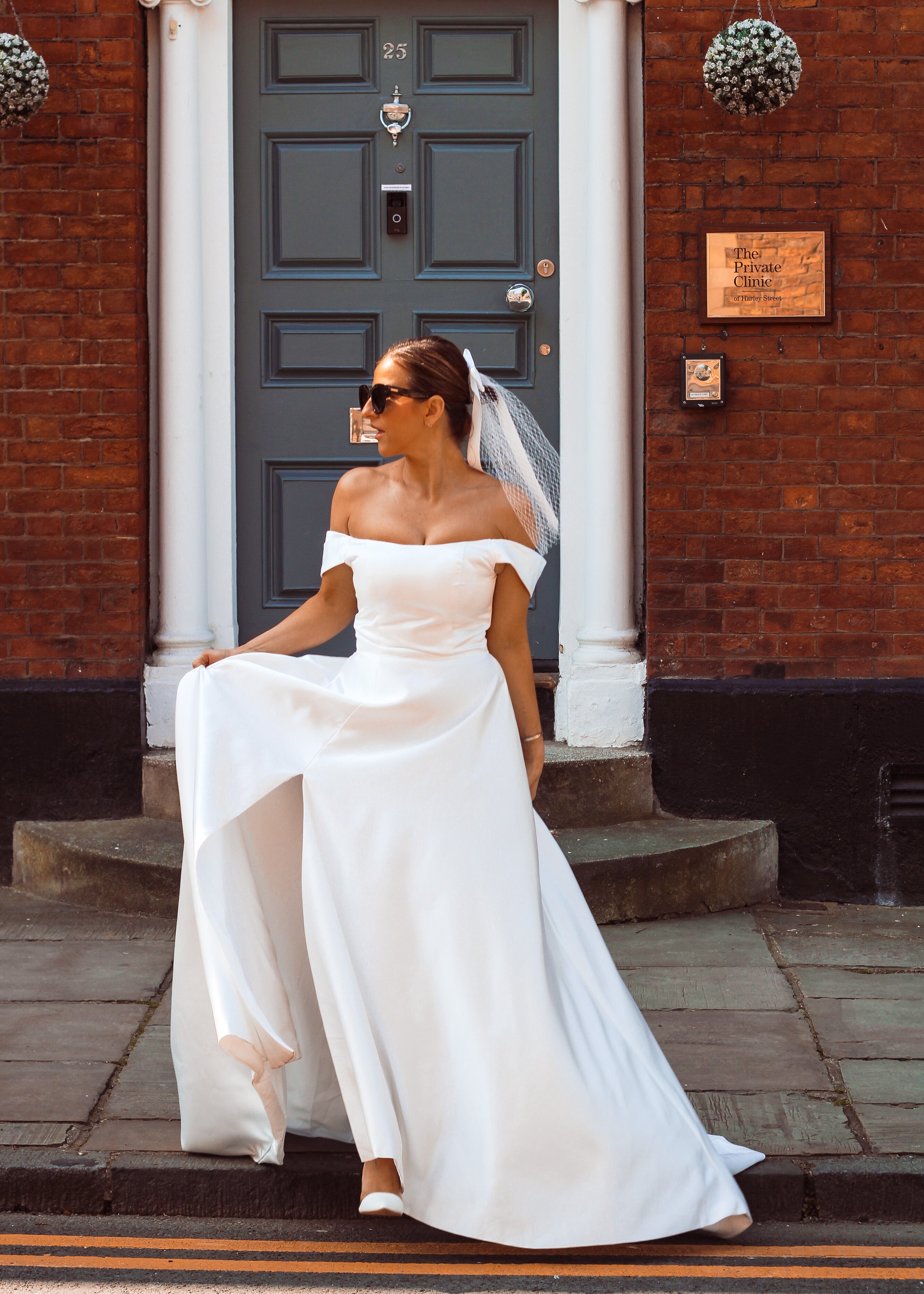 A young bride wearing Velo Bianco minimalistic off the shoulder satin wedding dress with buttoned back paired with short elegant wedding veil and white bridal heels. Manchester City hall wedding outfit for the modern bride. Wedding dress and black sunglasses outfit.