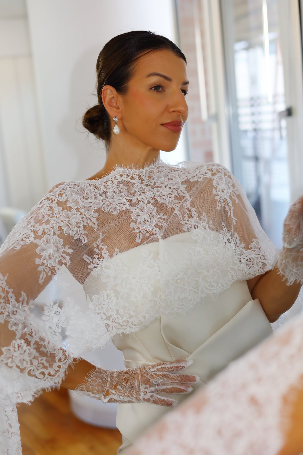 A woman wearing a white wedding dress with a sheer lace cape and matching lace gloves, standing indoors near a window.
