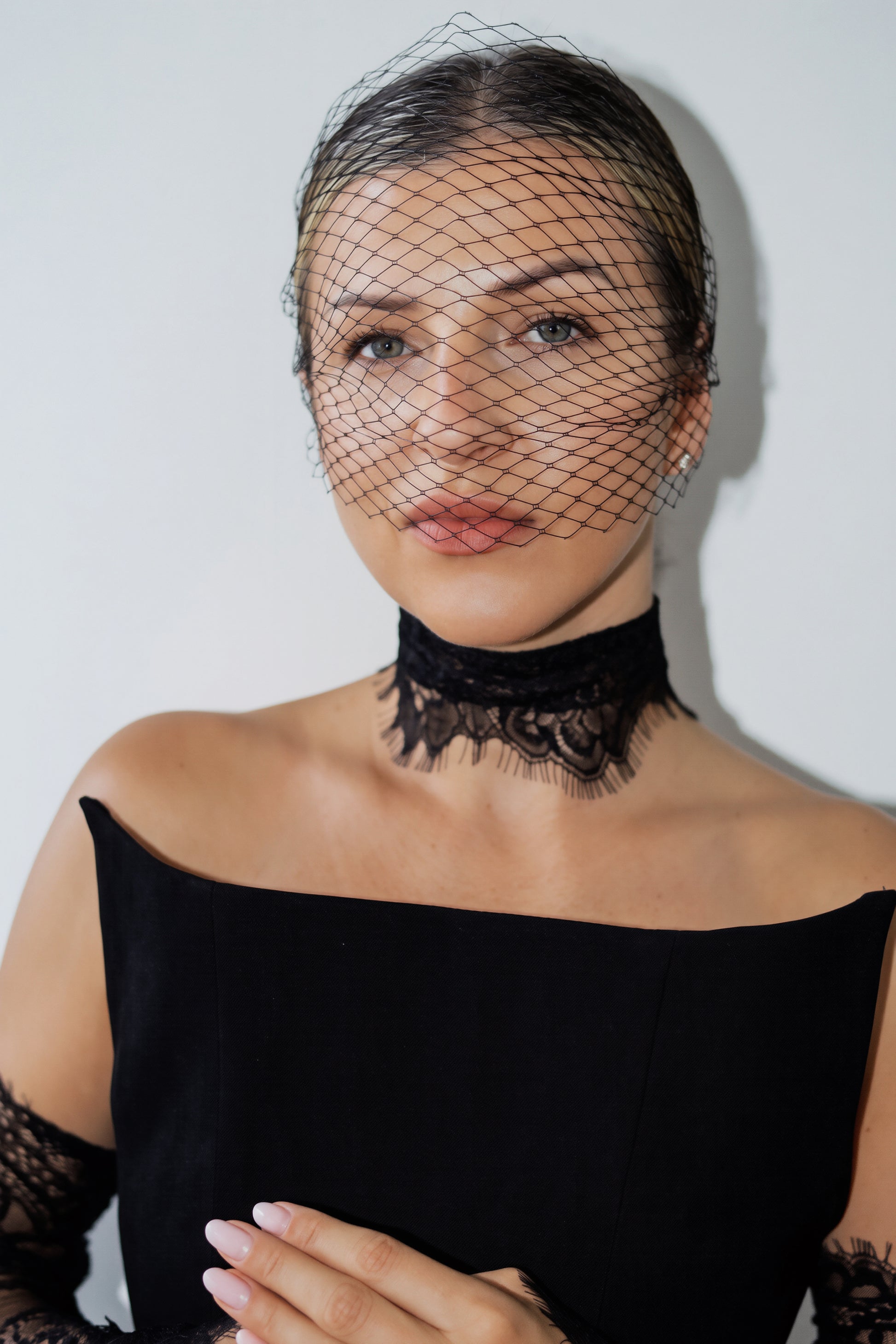 A woman wearing a black lace choker necklace with a matching black birdcage veil on her head, paired with a black off-the-shoulder top. She is posed against a plain light gray background.