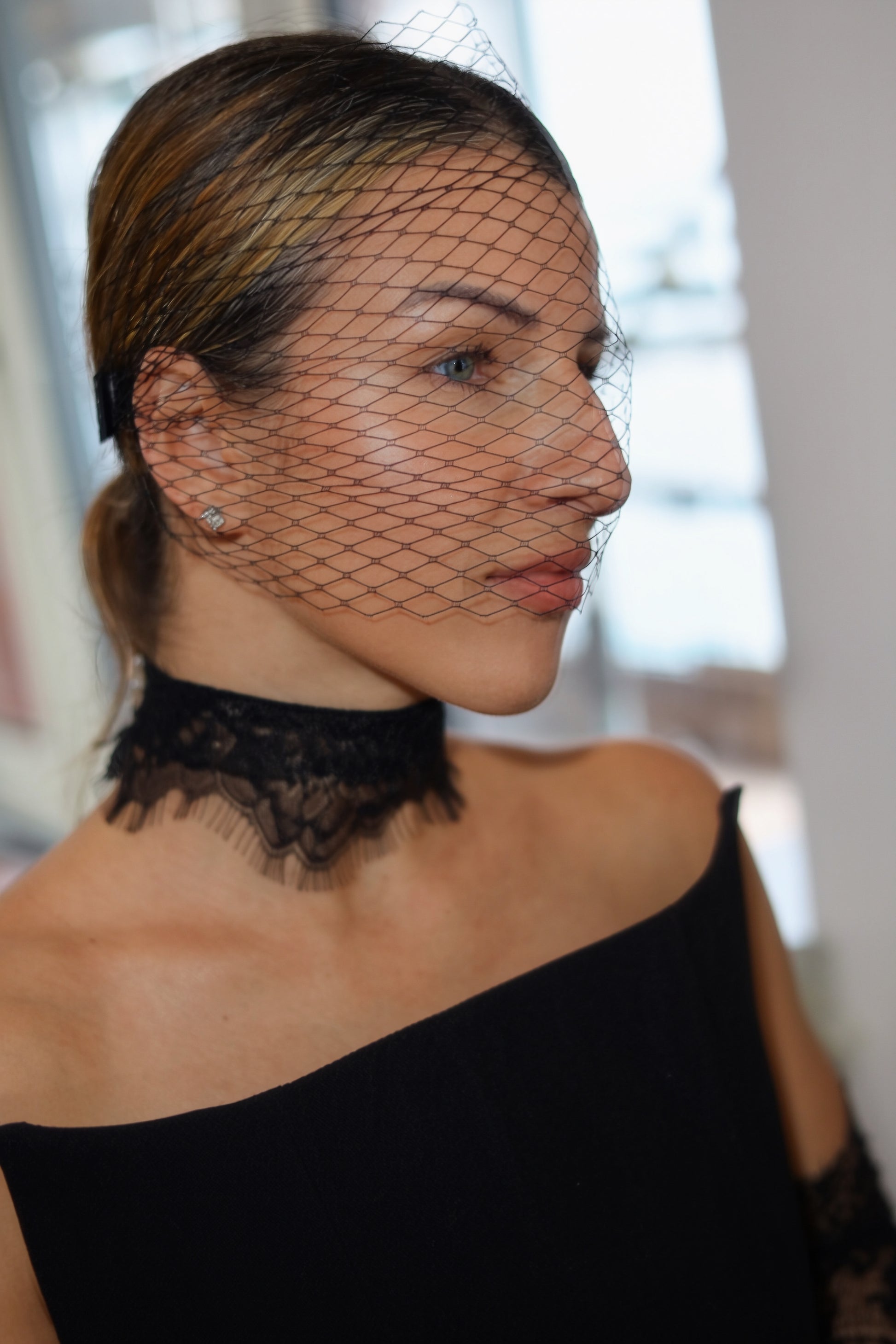 A woman in profile wearing a black lace choker and a black off-the-shoulder dress, with a black bird's-eye net veil draped over her face. She has blonde highlights in her hair and is wearing a small stud earring.