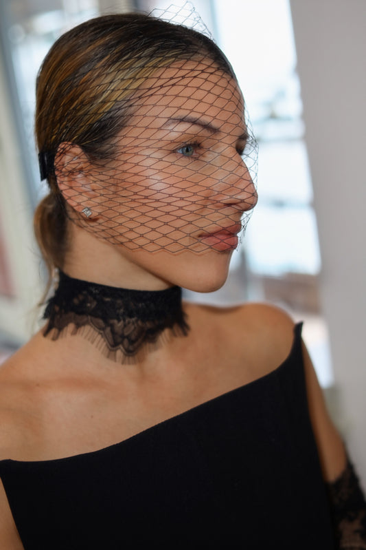 A woman in profile wearing a black lace choker and a black off-the-shoulder dress, with a black bird's-eye net veil draped over her face. She has blonde highlights in her hair and is wearing a small stud earring.
