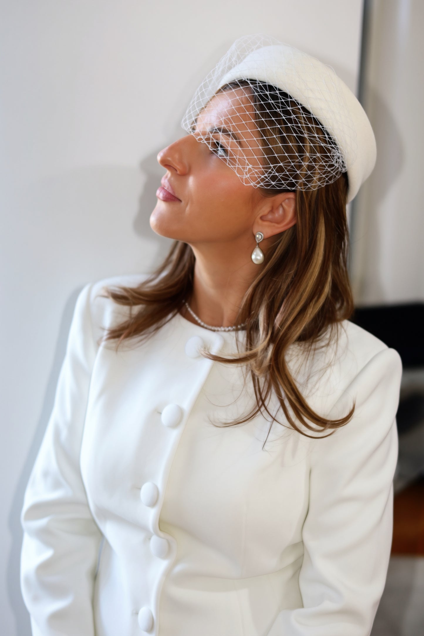 A woman wearing a white pillbox hat with a white net veil, a white jacket, and pearl earrings, looking upward against a plain light background.
