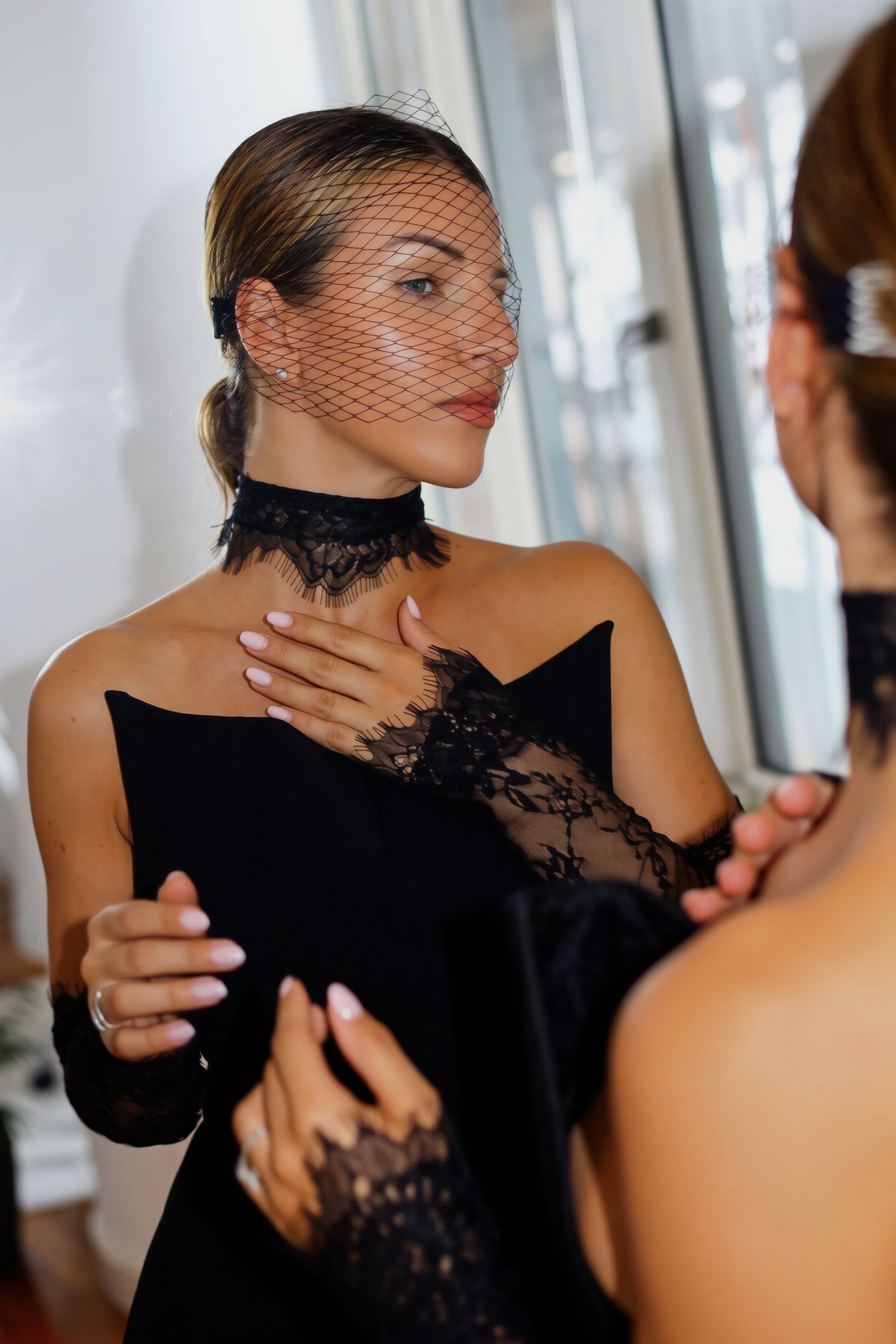 A woman wearing a black lace choker, matching lace gloves, and a black veil with a feather headpiece, seen in a mirror reflection while adjusting her dress.