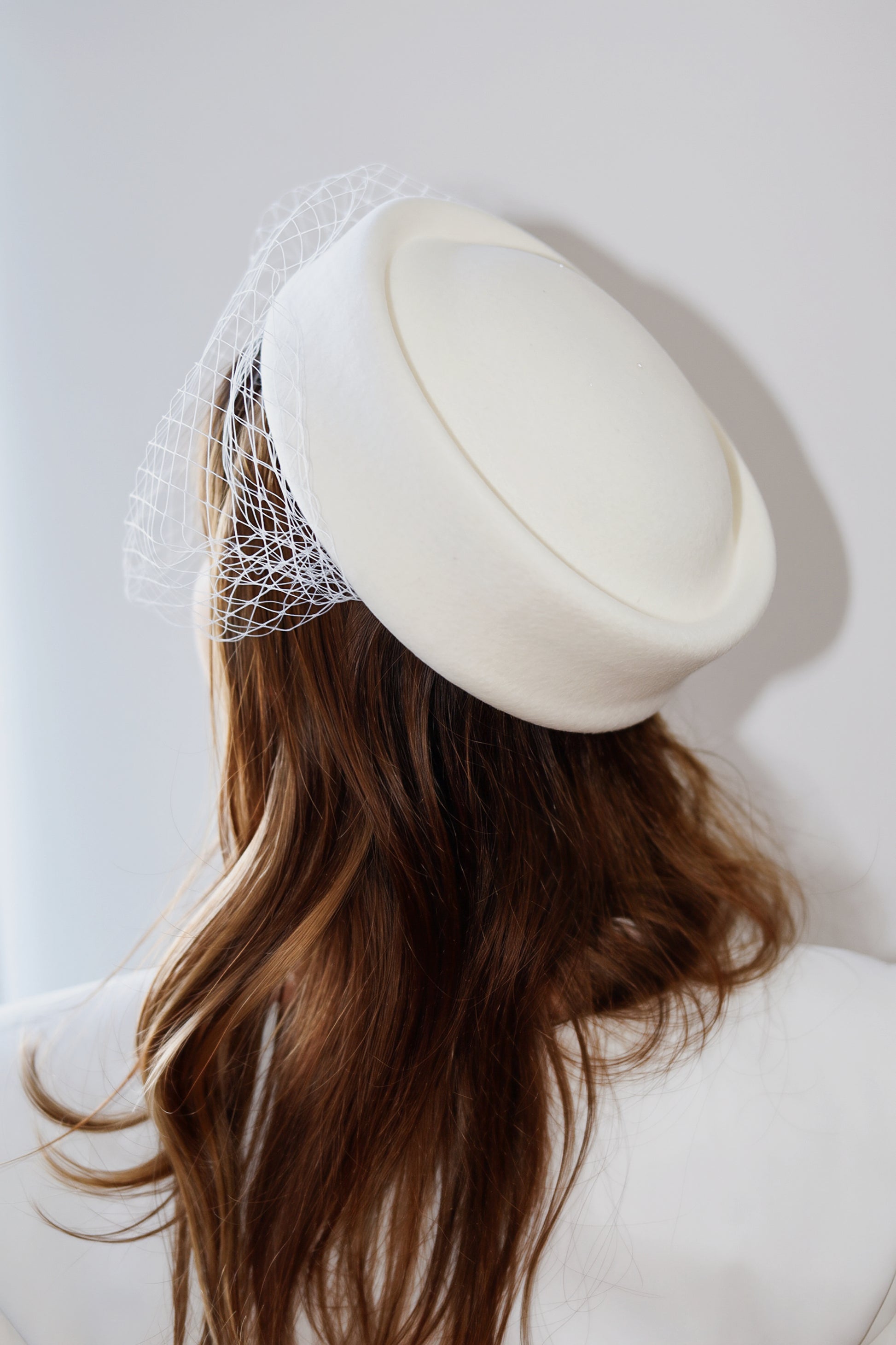 An off-white felt fascinator hat with a delicate white birdnet veil, shown on a model from the back against a light gray background.