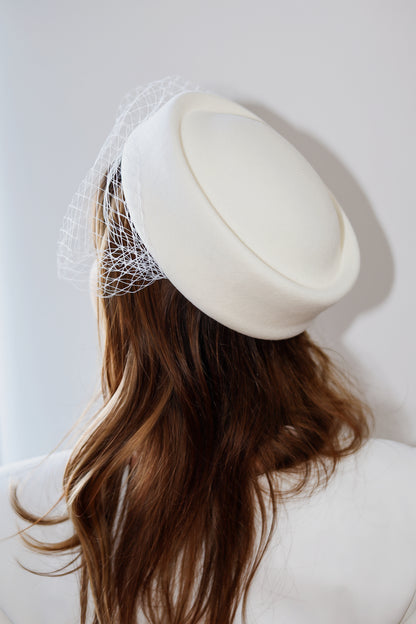 An off-white felt fascinator hat with a delicate white birdnet veil, shown on a model from the back against a light gray background.