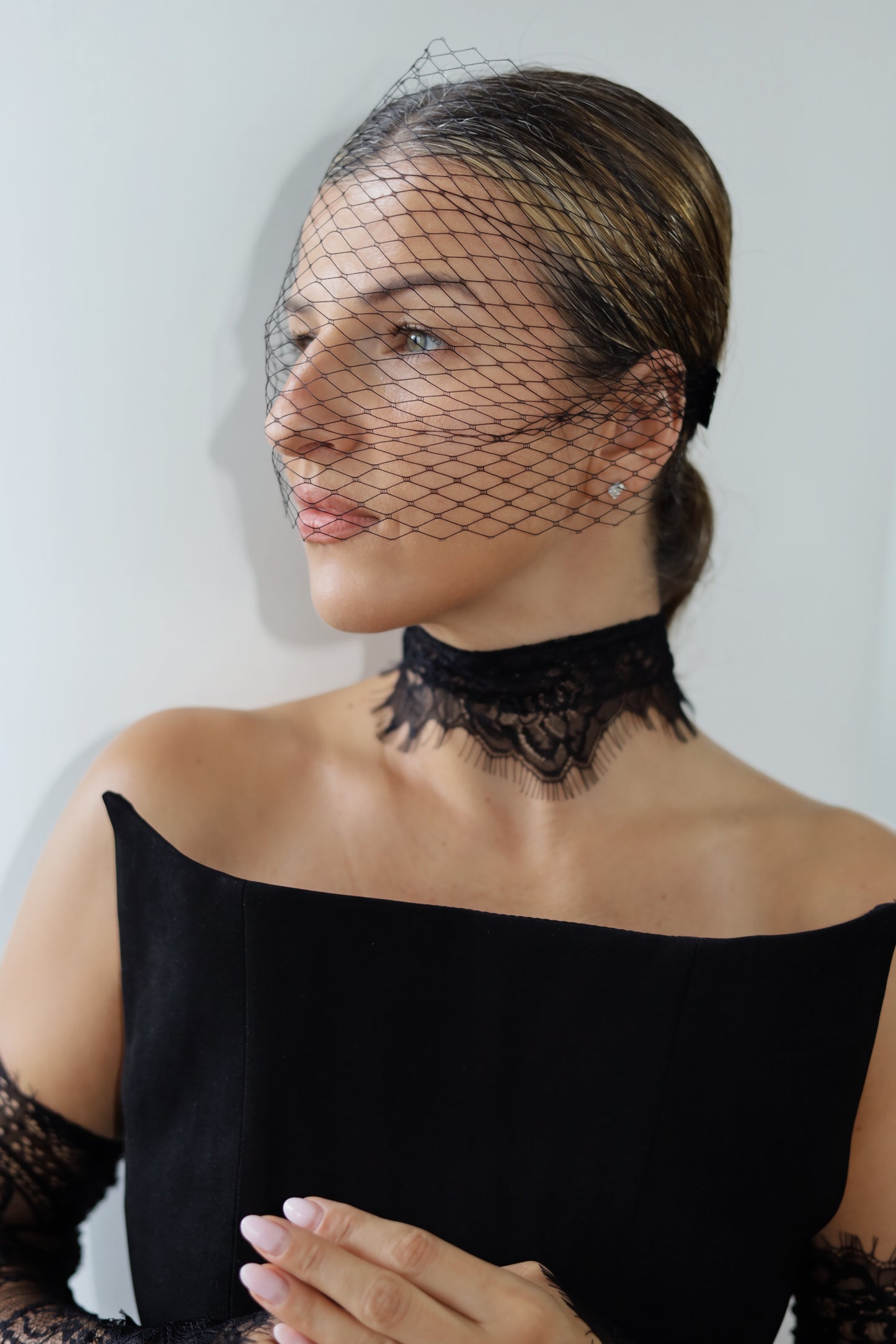 A woman wearing a black lace choker and a black bird's-eye net veil over her face, paired with a black off-the-shoulder top.