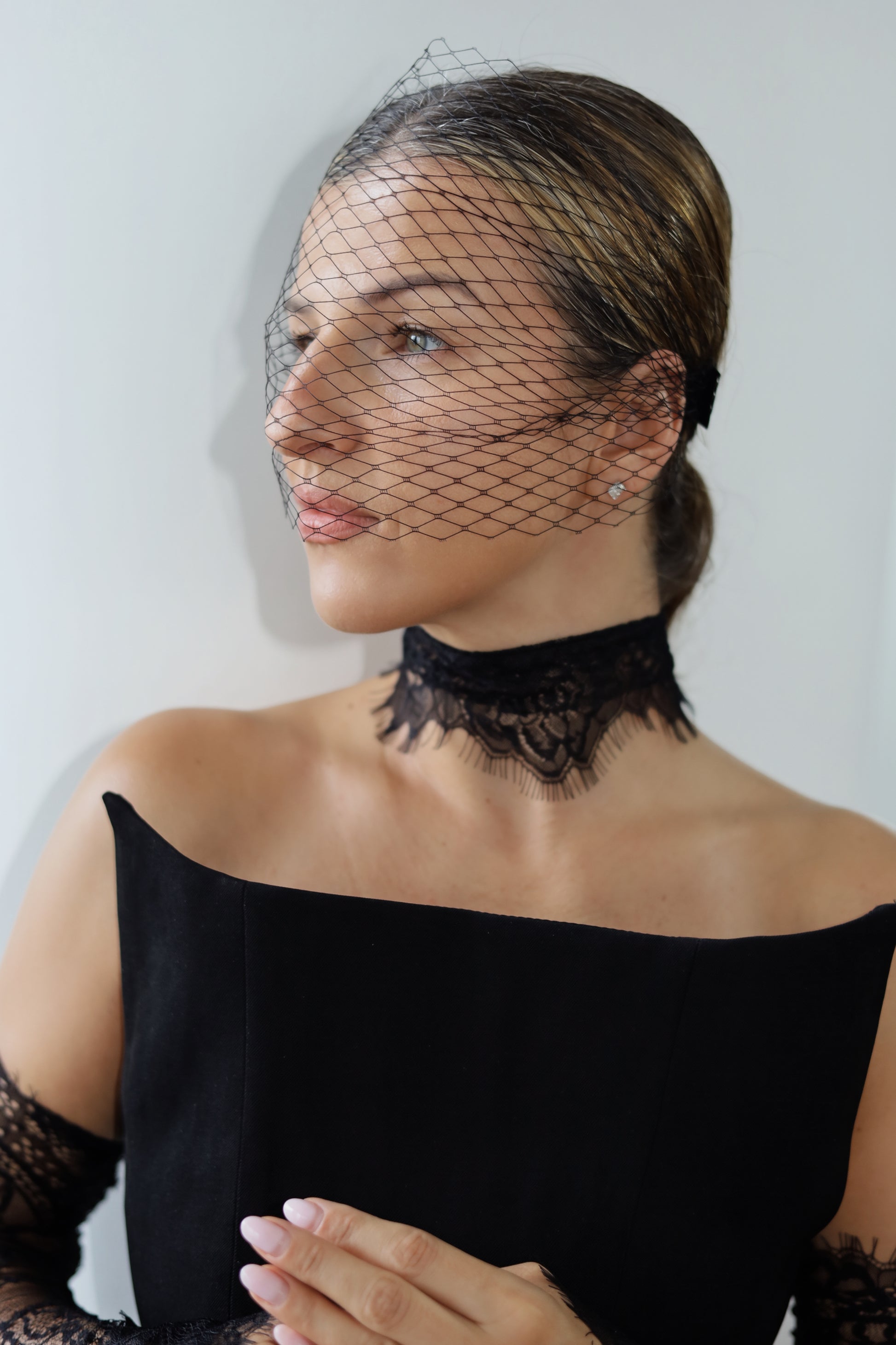 A woman wearing a black lace choker and a black bird's-eye net veil over her face, paired with a black off-the-shoulder top.