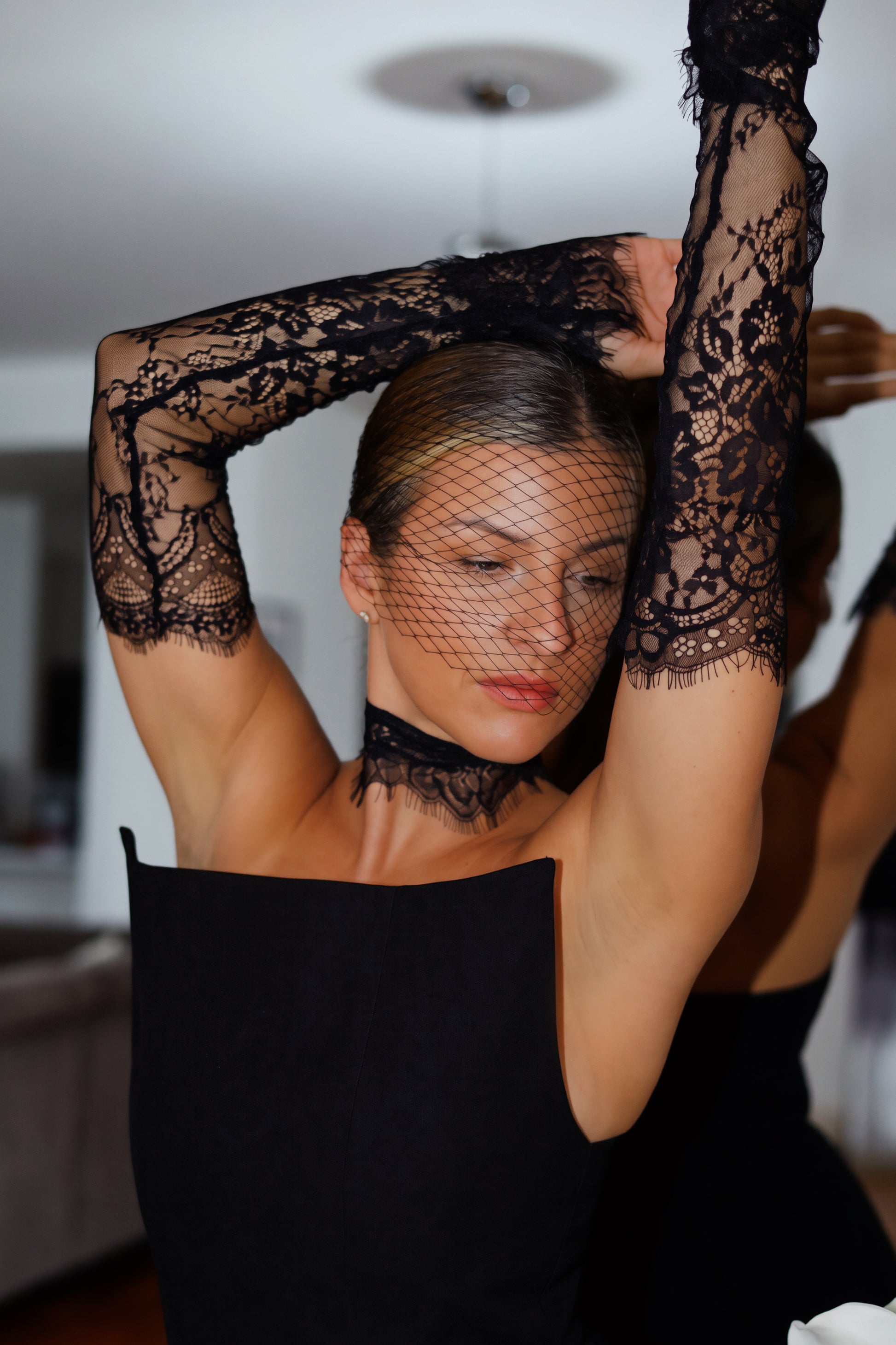 A woman wearing a black lace long-sleeve top with a matching black lace choker and fishnet head covering, posing with her arms raised in an indoor setting.
